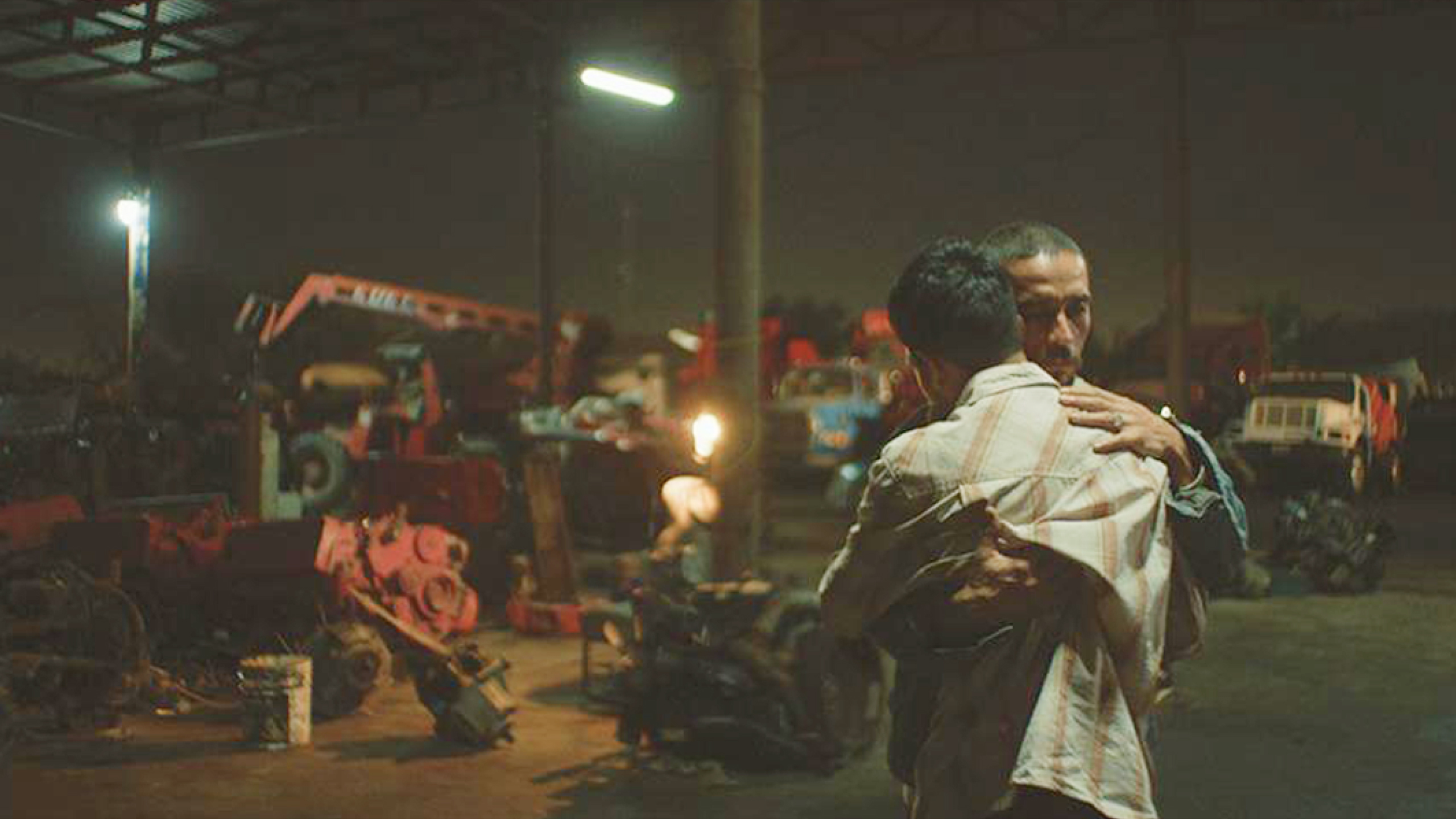 Due persone si abbracciano in un’officina all’aperto di notte, circondate da macchinari e luci artificiali.