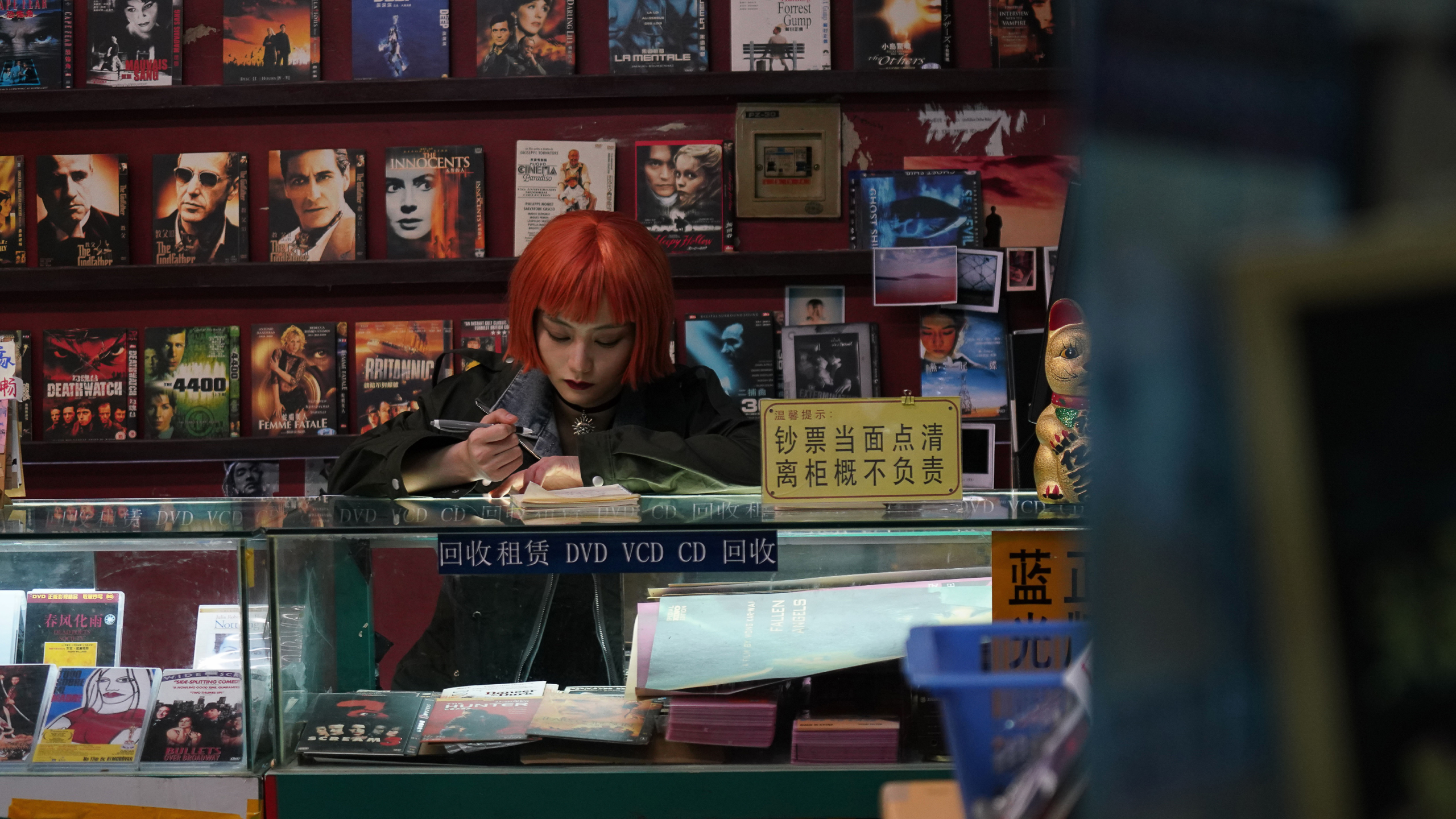 Una persona con parrucca rossa e trucco marcato scrive su un quaderno dietro il bancone di un negozio di DVD, circondata da locandine e copertine di film.