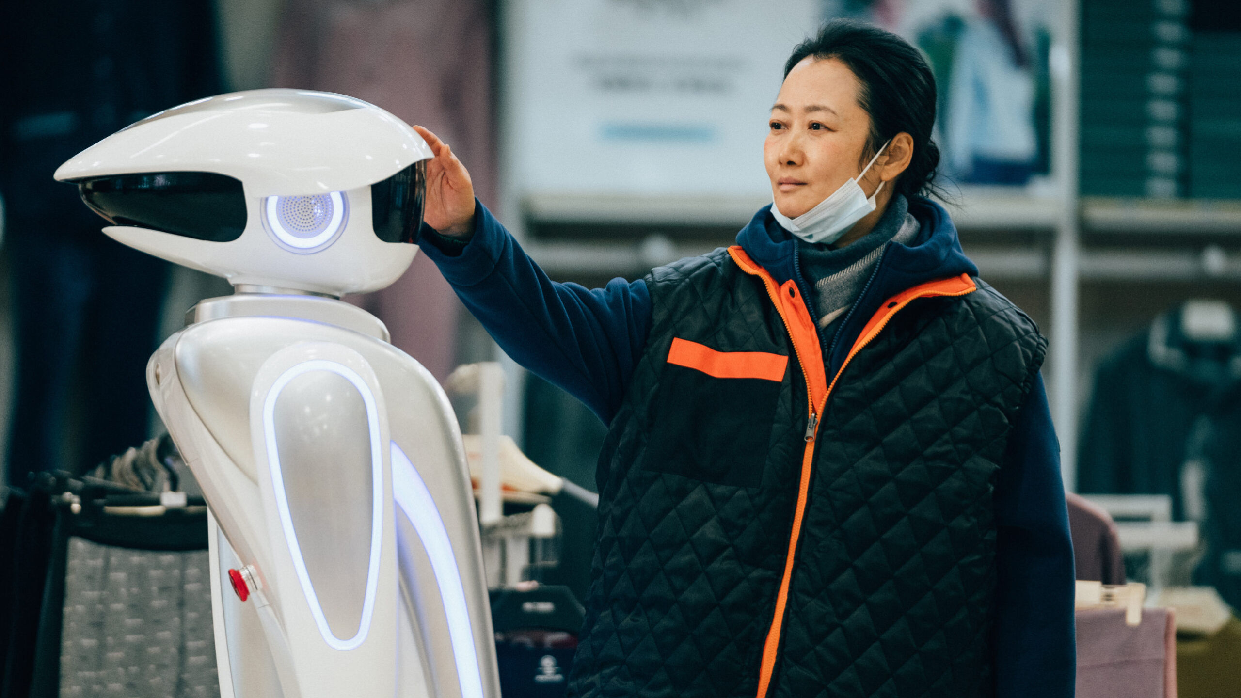 A woman touches the head of a white, futuristic-looking robot; she wears a dark jacket with orange details and a face mask lowered under her chin, while the robot has blue lights glowing on its body.