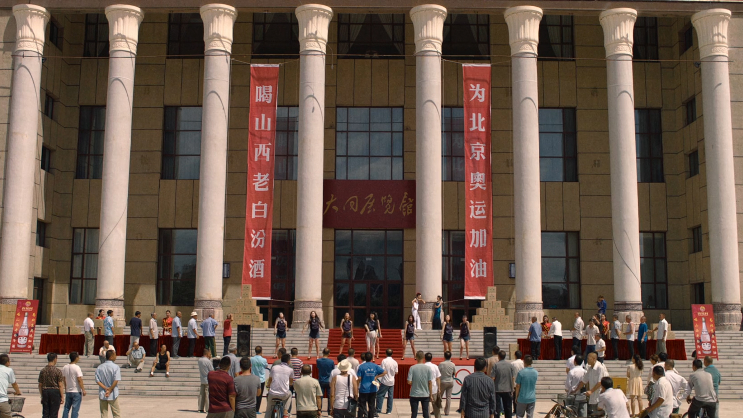 Ampia piazza davanti a un edificio monumentale con alte colonne bianche e striscioni rossi in caratteri cinesi; un gruppo di persone assiste a un’esibizione sul palco allestito sui gradini.