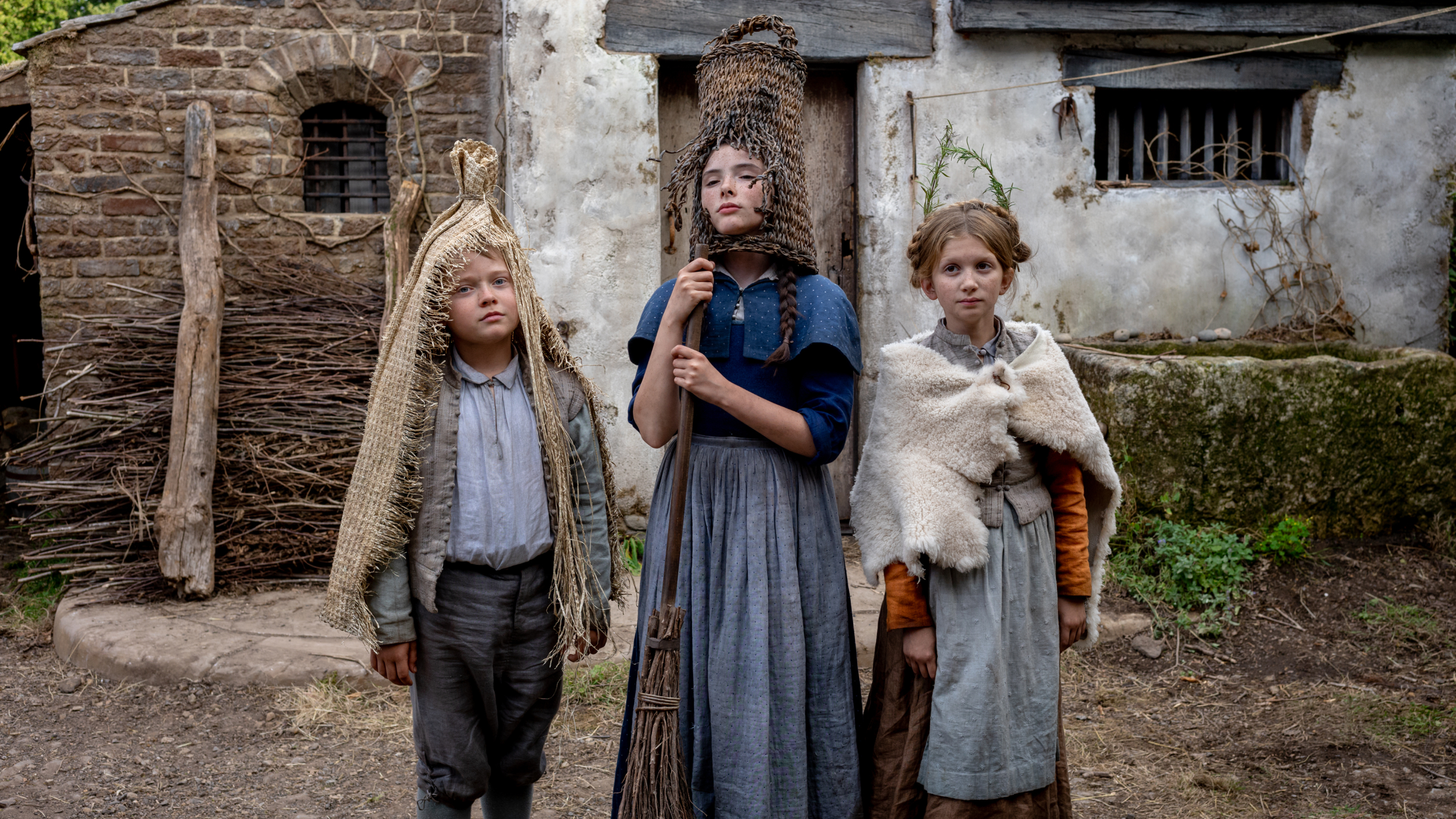 Three girls in period rural clothing stand in front of a rustic house; one wears a woven headpiece, another holds a broom, while the third is wrapped in a shawl.