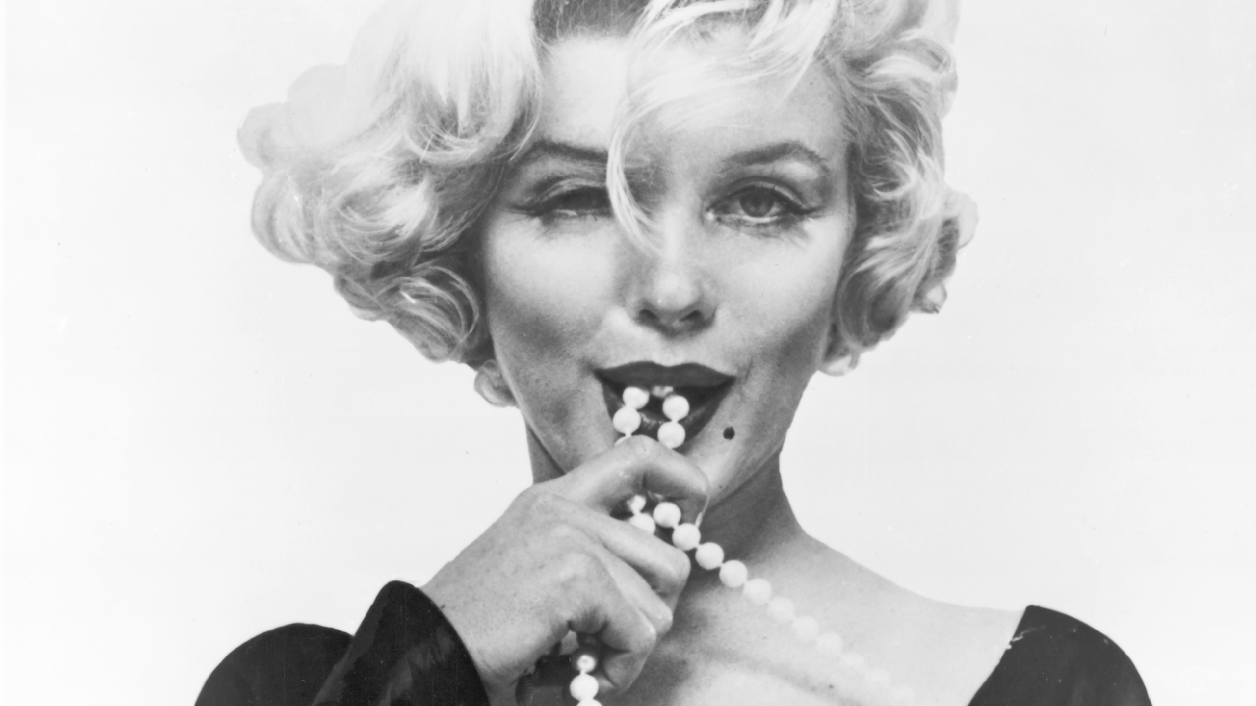 Ritratto fotografico in bianco e nero in primo piano di una persona con capelli corti ondulati, sguardo diretto verso l’obiettivo e un filo di perle portato vicino alla bocca.